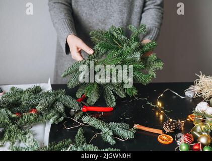 Junge Frau in warmen grauen Strickkleid macht diy Tanne weihnachtskranz in der Hand auf dem Tisch zu Hause Stockfoto