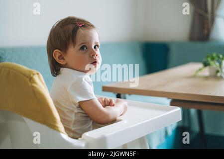 Charmantes kleines Mädchen sitzt in Hochstuhl für die Fütterung in der hellen Küche zu Hause Stockfoto