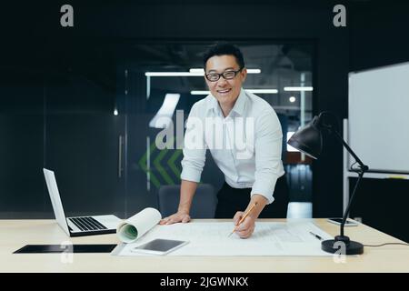 Porträt eines erfolgreichen und erfahrenen asiatischen Architekten, Mann, der mit einer Planzeichnung über den Tisch steht, lächelt und die Kamera anschaut, der im modernen Designstudio arbeitet Stockfoto