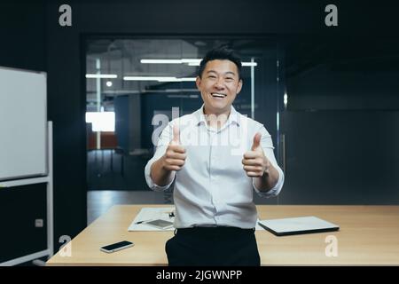 Porträt eines erfolgreichen asiatischen Geschäftsmanns-Chefs, eines Mannes, der im Büro neben dem Tisch steht und die Kamera anschaut und lächelt und mit den Händen den Daumen nach oben zeigt Stockfoto