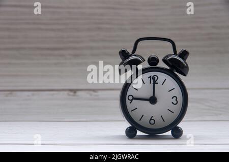 Schwarzer Wecker, isoliert auf Holzabdeckung. Die Uhr wurde auf 9 Uhr eingestellt. Speicherplatz kopieren. Stockfoto