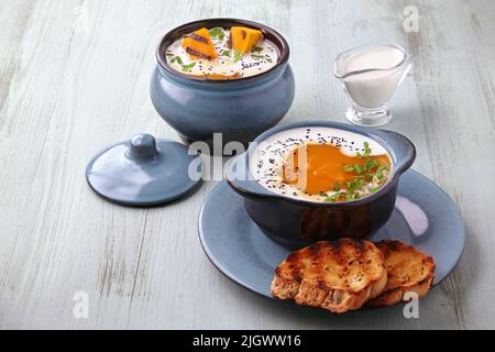 Kürbis-Herbstpüree-Suppe mit Kräutern, Samen und Rahm in einer Schüssel und Topf mit gerösteten Brotscheiben und Kürbiskernen auf einem blauen Holztisch. Vorderansicht Stockfoto