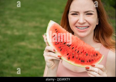Schöne rothaarige Frau, die mit Zahnspangen lächelt und im Sommer ein Stück Wassermelone im Freien essen will Stockfoto