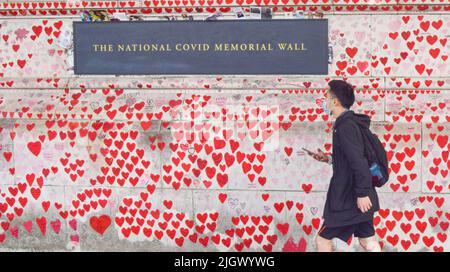 London, Großbritannien. 13.. Juli 2022. Ein Mann mit einer schützenden Gesichtsmaske geht an der National Covid Memorial Wall vor dem St. Thomas' Hospital vorbei, während die britischen Covid-Todesfälle über 200.000 liegen. Tausende rote Herzen wurden an die Wand gegenüber dem Parlament gemalt, eines für jedes Leben, das durch das Coronavirus verloren wurde. Kredit: Vuk Valcic/Alamy Live Nachrichten Stockfoto