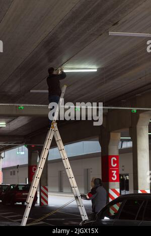 Stadtarbeiter Elektriker repariert LCD-Lampe auf Mall Parkplatz. Der zweite Arbeiter unterstützt ihn. Beruf, der Komfort bietet, Strom in Städten. Stockfoto