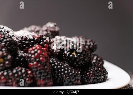 Brombeeren bestreut mit Zucker auf einem Teller auf schwarzem Hintergrund, Nahaufnahme von der Seite. Stockfoto