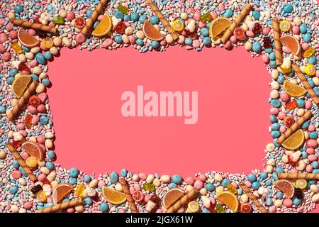 Mehrfarbige glasierte Dragees und andere Süßigkeiten sind in Form eines Rahmens auf einem korallenen Hintergrund angeordnet. Stockfoto