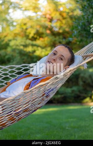 Vertikales Porträt eines jungen, gutaussehenden Mannes, der im Sommer in einer Hängematte im Hausgarten vor die Kamera schaut Stockfoto