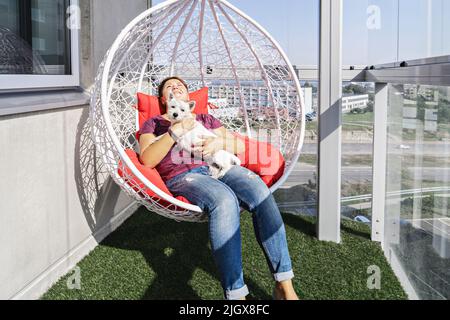 Millennial Frau mit weißem Welpen sitzt in einem modernen Hängesessel auf dem Balkon des Wohnhauses und lächelt Stockfoto