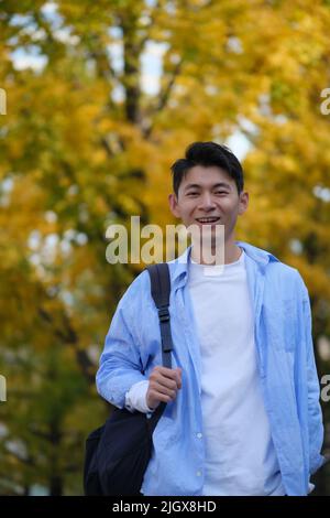Asiatischer männlicher Student und verschwommener gelber Ginkgo-Biloba-Baum im Herbst. Eine Schultertasche tragen und vor der Kamera lächeln Stockfoto