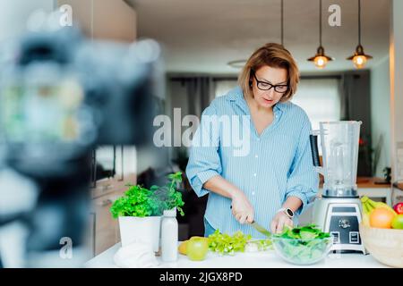 Frau im mittleren Alter bloggt für ihren Wellness-Lifestyle-Kanal über ein gesundes, ausgewogenes Leben in der Küche ihres Hauses. Erstellen von Videoinhalten für s Stockfoto
