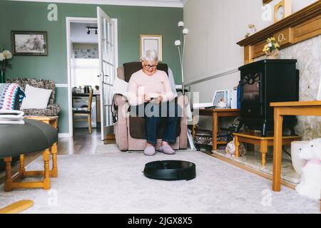 Ältere Frau, die einen Roboter-Staubsauger über Telefoneinstellungen verwendet, während sie zu Hause auf einem Sessel sitzt. Ältere Dame mit Mobiltelefon und Kontrolle Stockfoto