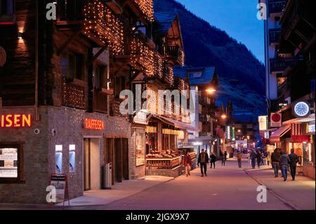 ZERMATT, SCHWEIZ - APR 11, 2022: Touristen gehen und besichtigen auf einer Straße in der Stadt Zermatt bei Sonnenuntergang. Stockfoto
