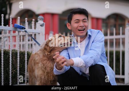 Asiatischer junger Mann und sein verspielter goldener Retriever-Hund lächeln im öffentlichen Park vor der Kamera Stockfoto