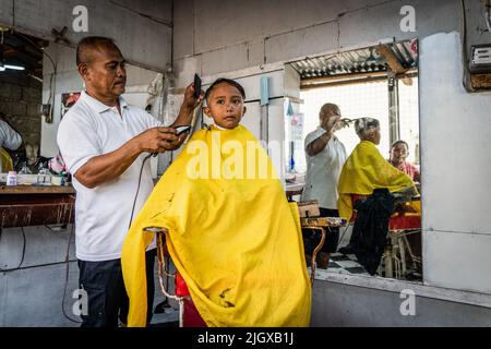 Moalboal, Philippinen. 13.. Juli 2022. Ein Junge bekommt seine Haare in einem Friseurladen an einer geschäftigen öffentlichen Bushaltestelle geschnitten. Tägliches Leben im Moalboal District, Cebu, Philippinen. Das Gebiet, das einst für sein Tauchen und seine massiven Sardinenwolken bekannt war, hat sich schwer getan, den Tourismus wieder zu beleben, auch nachdem die Reisebeschränkungen von Covid-19 aufgehoben wurden, da die Infrastruktur durch den Taifun Rai, auch bekannt als Super Typhoon Odette, beschädigt wurde, was einen Großteil des Gebiets dezimiert und mit dem Wiederaufbau zu kämpfen hat. Kredit: SOPA Images Limited/Alamy Live Nachrichten Stockfoto