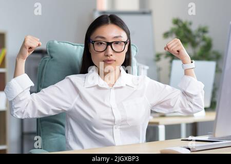 Porträt einer jungen, schönen asiatischen Geschäftsfrau, die mit ihren eigenen Händen zeigt, dass sie stark und erfolgreich ist. Sitzt am Schreibtisch im Büro in einem weißen Hemd und einer Brille. Stockfoto