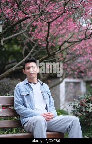 Ein hübscher, lächelnder ostasiatischer junger Mann, der im Frühjahr auf einer Bank unter einem Blütenbaum mit rosa Pflaumen saß und in die Kamera schaute Stockfoto