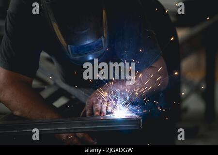 Der Handwerker, der an seinem Arbeitsplatz in der Werkstatt Schweißen und Schleifen durchführt, während die Funken um ihn herum „fliegen“. Er trägt einen Schutzhelm und Stockfoto