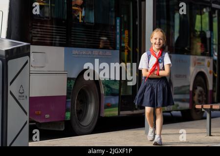 Huzhou, China 22 June 2021: Glückliches kaukasisches Mädchen kehrt in Uniform und roter Krawatte bei Pioneer in die Schule zurück. Am Bahnhof wartet ein Kind auf einen öffentlichen Bus Stockfoto