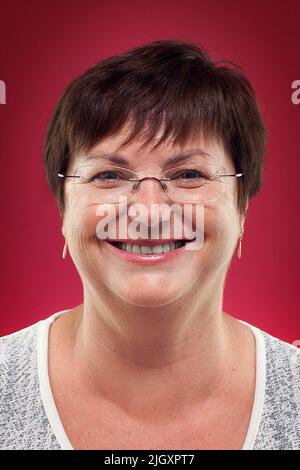 Porträt einer Frau mittleren Alters, die eine Brille und eine Kassualkleidung trägt und vor einem roten Hintergrund auf die Kamera blickt Stockfoto