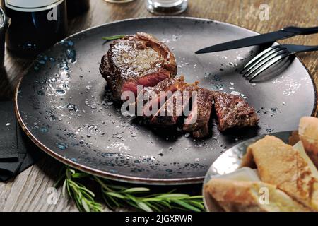 Nahaufnahme geschnittenes mittelseltenes Steak, serviert auf Teller mit Besteck und Schüssel mit frisch gebackenem Brot. Hauptgericht, Restaurantgericht, Esskonzept Stockfoto