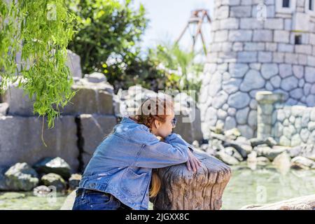 Eine Frau, die unter Weide sitzt, blickt auf den Fluss in der Nähe des Steinschlosses im Vergnügungspark. Frühlingsruhe. Parkdekorationen. Selektiver Fokus. Stockfoto