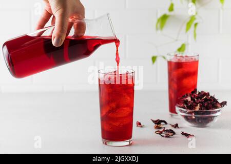 Roter Kräutertee aus Hibiskusblättern wird aus einer Kanne mit Eis in ein Glas gegossen. Stockfoto