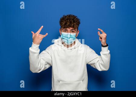 Junger Mann mit lockigen Haaren, der eine Gesichtsmaske für Coronavirus trägt, wodurch die Felsgesture auf blauem Studiohintergrund isoliert wird Stockfoto