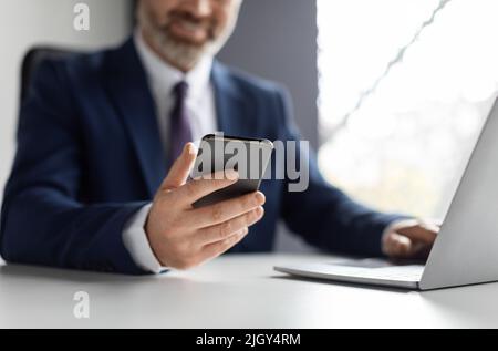 Lächelnd Reife Geschäftsmann Mit Smartphone Und Die Arbeit An Laptop In Modern Office Stockfoto