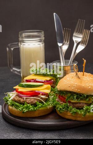 Cheeseburger mit Grüns auf Brötchen mit Sesam. Glaskrug mit Bier. Messer und Gabel in Zinn. Draufsicht. Schwarzer Hintergrund Stockfoto