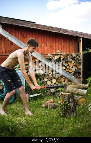 Ein dünner, drahtiger Teenager, barfuß, in schwarzen Shorts, hackt an einem klaren, sonnigen Sommertag mit einer Axt auf einem Stumpf, auf grünem Gras Holz Stockfoto