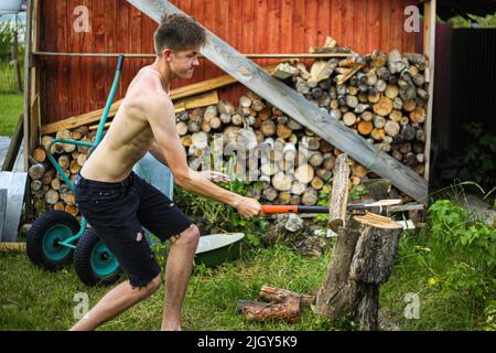 Ein dünner, drahtiger Teenager, barfuß, in schwarzen Shorts, hackt an einem klaren, sonnigen Sommertag mit einer Axt auf einem Stumpf, auf grünem Gras Holz Stockfoto