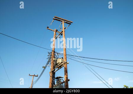 Externer Transformator auf Holzmasten, die mit elektrischer Energie auf blauem Himmel verbunden sind Stockfoto