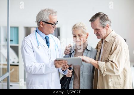 Seriöse, gut aussehende Ärztin mit Stethoskop am Hals und digitalem Tablet, während sie mit ihrem Mann nützliche Informationen zur Genesung der Patientin gibt Stockfoto