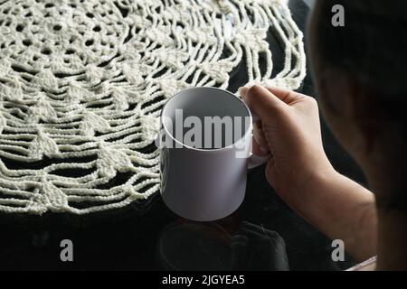 Detailaufnahme eines Latina-Mädchens, das eine Kaffeetasse in der Hand hält und an einem Glastisch mit einem weißen gehäkelten Mittelteil sitzt. Tischdecke aus Wolle gewebt Stockfoto