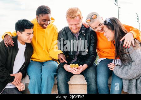 Fünf junge Freunde sehen sich ein Video auf einem Mobiltelefon an und lächeln zusammen. Stockfoto