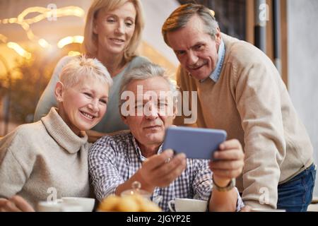 Hübscher älterer Mann im karierten Hemd, der am Tisch sitzt und das Smartphone hält, während er Videos mit Freunden teilt Stockfoto