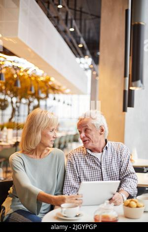 Inhalt modernes Seniorenpaar in lässigen Outfits, das im Restaurant am Tisch sitzt und Tablet benutzt, während es im Online-Shop Dinge auswählt Stockfoto