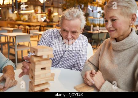 Eine Gruppe konzentrierter älterer Freunde, die im Freizeitzimmer des Pflegeheims am Tisch sitzen und Jenga spielen Stockfoto