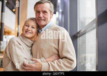 Porträt eines glücklichen, schönen Seniorenpaares in beigen Pullovern, die am Fenster stehen und sich gegenseitig kuscheln Stockfoto