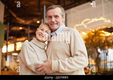 Porträt eines glücklichen verheirateten älteren Paares in lässigen Pullovern, die sich in einem modernen Restaurant voller Lichter umarmen Stockfoto