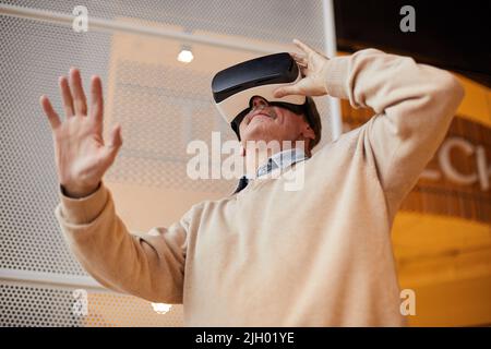 Unten ein neugieriger älterer Mann mit Schnurrbart, der die Hand sticht, während er eine Virtual-Reality-Brille verwendet Stockfoto
