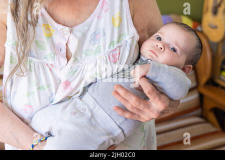 Nahaufnahme eines entzückenden, zwei Monate alten Mädchens, das einen grauen Pullover trägt, der in die Arme der Großmutter gewickelt ist und den Finger ergreifend. Mit Kopierbereich. Stockfoto