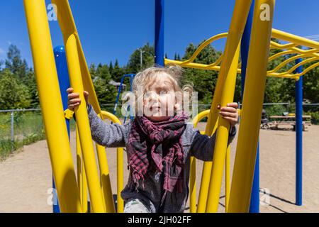 Nahaufnahme eines gesunden, zweijährigen Jungen mit blondem, welligem Haar, mit einem Klettergerüst auf einem Kinderspielplatz mit verschwommenem Gerät im Hintergrund. Stockfoto