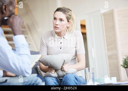 Wütende junge Frau in zwanglosem Outfit sitzt auf dem Sofa und drückt sich auf das Kissen, während sie bei der Psychologiesitzung Emotionen zum Ausdruck bringt Stockfoto