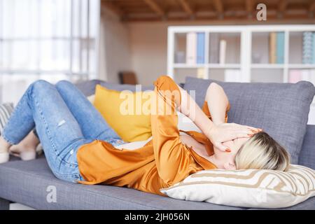 Verstörte junge Frau in orangefarbener Bluse, die bei psychoanalytischer Therapie auf dem Sofa liegt und beim Weinen das Gesicht mit den Händen bedeckt Stockfoto