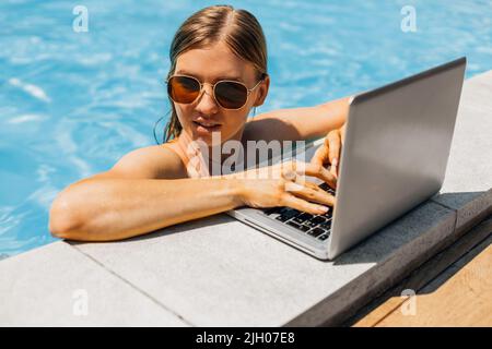 Eine wunderschöne Frau, die mit einem Laptop aus der Ferne arbeitet, sitzt in einem Pool in einem tropischen Spa. Freiberuflicher Unternehmer. Freiberufliche Frau im Urlaub, concep Stockfoto