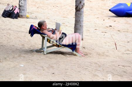 Eine Frau sitzt in einem Liegestuhl und liest ein Buch. Tägliches Leben am Strand in Jomtien, Pattaya, Thailand, Asien. Stockfoto