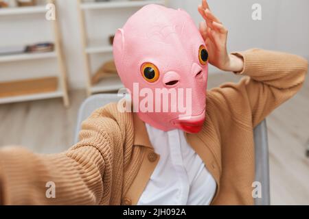 Lustige blonde Dame in rosa Fischmaske warmen Pullover tun Selfie verwenden Telefon sitzen im Sessel im modernen Zuhause Interieur. Pause von der Arbeit, Pause machen Stockfoto