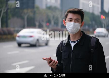 Asiatischer junger Mann mit Gesichtsmaske, der das Telefon hält, um ein Taxi in der Nähe der Straße zu rufen, und in die Kamera schaut Stockfoto